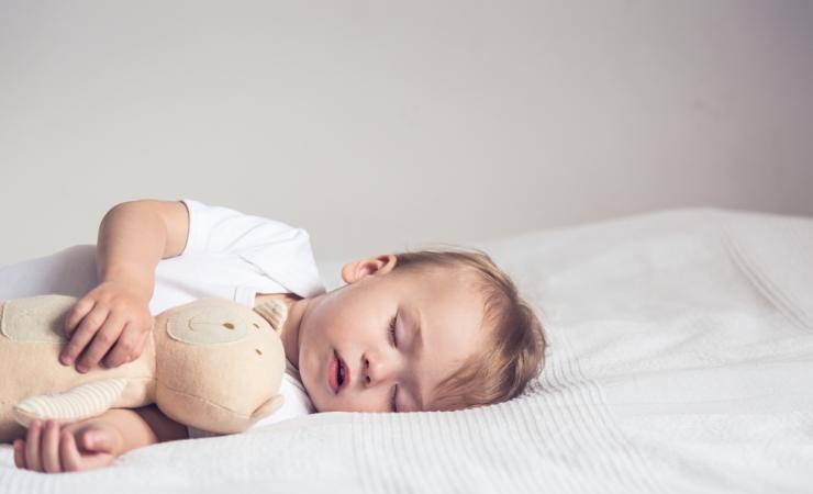 A baby is lying sleeping on its left side on a bed. The baby is cuddling a beige teddy bear.