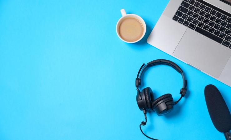 A laptop keyboard, headphones, microphone and a cup of coffee are viewed from above. The background is bright blue. Image by Boiarkina Marina via Shutterstock.