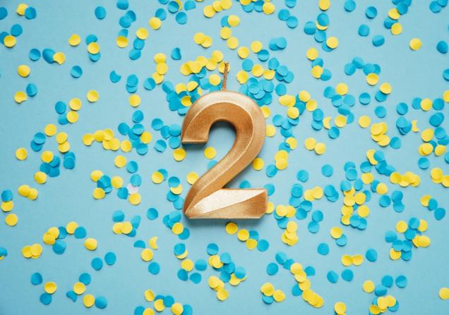 A golden candle in the shape of the number 2 on a blue background covered with blue and yellow confetti. Image by Serenko Natalia via Shutterstock. 