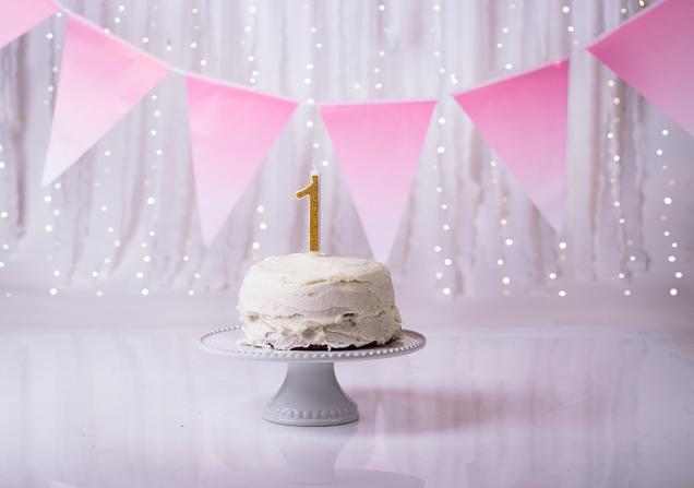 A white cake on a white stand with a gold candle in the shape of the figure '1'. There is pink bunting in the background. Image by Nexx via Pexels