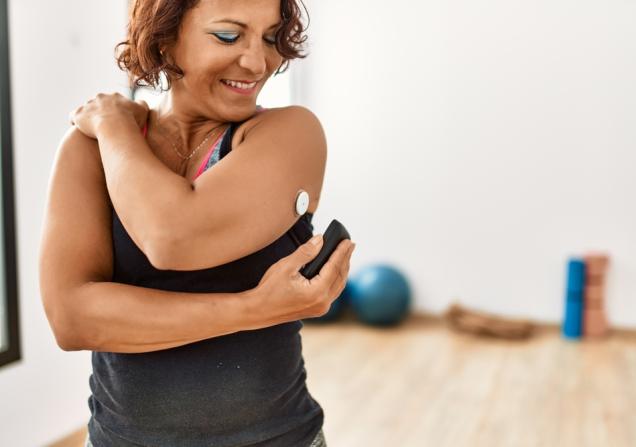 In the foreground of the picture is a woman with diabetes checking her glucose levels by scanning a glucose monitor attached to her upper arm. She is wearing sports clothes and in the background, out of focus, is some exercise equipment. Image by Krakenimages.com via Shutterstock