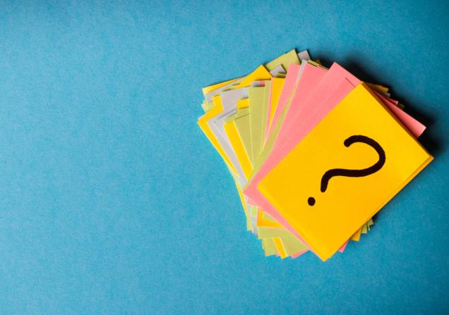 A pile of pieces of yellow, green, blue and pink paper against a blue background. The top piece of paper is bright yellow and has a question mark on it. Image by SNeG17 via Shutterstock.
