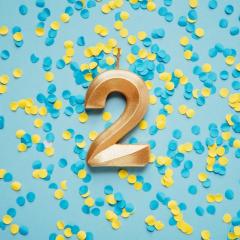 A golden candle in the shape of the number 2 on a blue background covered with blue and yellow confetti. Image by Serenko Natalia via Shutterstock.