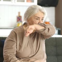 A senior woman coughing into her elbow. She has white skin and long white hair tied back. She's wearing a beige long-sleaved top and sitting on a grey sofa. Image by Red Stock via Shutterstock.