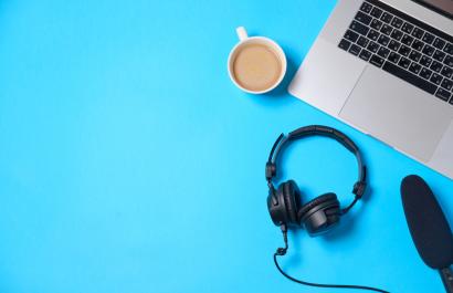 A laptop keyboard, headphones, microphone and a cup of coffee are viewed from above. The background is bright blue. Image by Boiarkina Marina via Shutterstock.