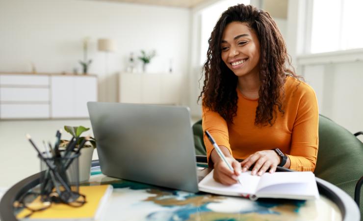 Woman sitting at desk working on laptop taking notes. Image by Prostock-studio via Shutterstock.