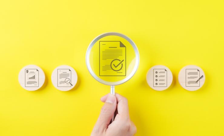 In the centre of the image, a hand is holding a magnifying glass over an icon of a document with a tick mark on it. On either side are more icons with documents and tick marks on wooden circles. The background of the image is bright yellow. Image by SuPatMaN via Shutterstock.