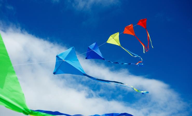 A string of coloured kites flying high against a blue sky with a few white clouds. Image by Sergey Novikov via Shutterstock.