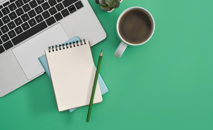 A laptop, spiral-bound notepad, pencil, cup of coffee and a small plant are seen from above. The background is green. Image by Prathankarnpap via Shutterstock.