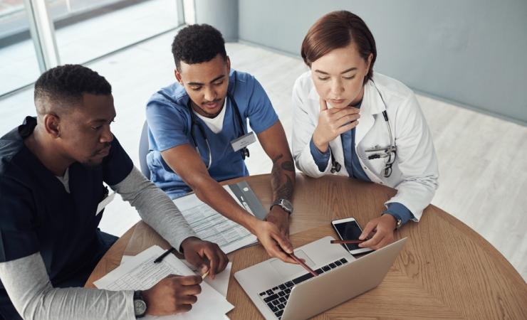 Three people sitting round a table looking at a laptop. Two of them have stethoscopes around their neck. Image by PeopleImages.com - Yuri A via Shutterstock.
