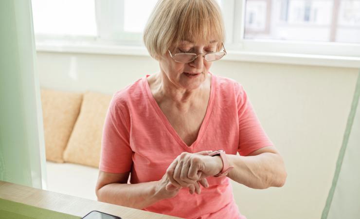 A senior woman is looking at a fitness tracker on her wrist. She has short blonde hair and glasses and is wearing a pink t-shirt. Image by Maria Symchych via Shutterstock.