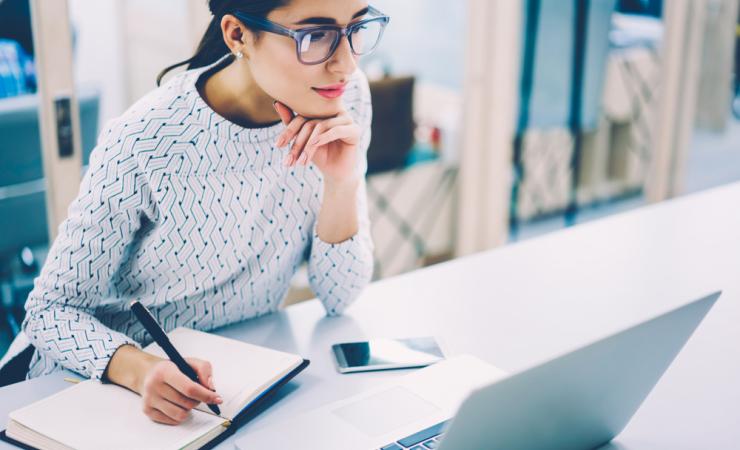 A woman looking at a computer screen and taking notes. 