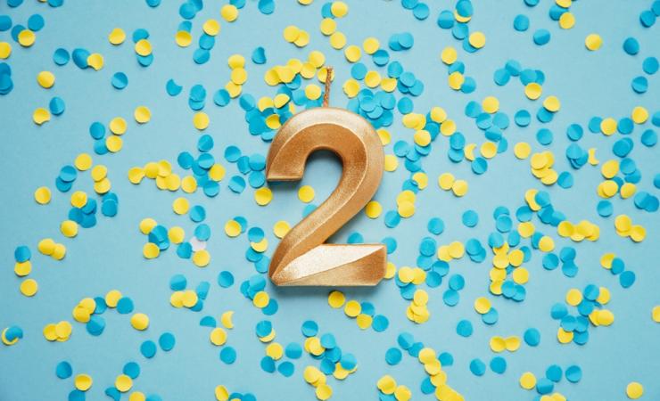 A golden candle in the shape of the number 2 on a blue background covered with blue and yellow confetti. Image by Serenko Natalia via Shutterstock.