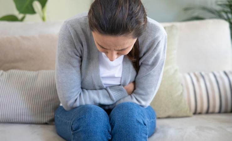 A woman is sitting on a sofa, facing towards us. She's doubled over in pain, so we can't really see her face, just the top of her head. She's wearing a white top, grey cardigan and blue jeans. Her long dark hair is tied back in a ponytail.