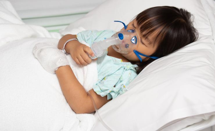 A young girl lying in a hospital bed with her eyes closed and an oxygen mask on. Image by Huttsu via Shutterstock.