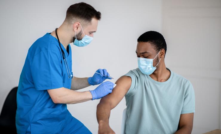 A doctor in blue scrubs gives a man in a pale turquoise t-shirt his COVID-19 vaccination. They're both wearing facemasks. Image by Prostock-studio via Shutterstock