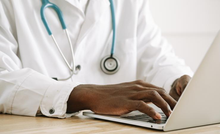 A close-up of a doctor typing at a computer. The doctor is wearing a white doctor's coat and a stethoscope. Image by Ivan Samkov via Pexels.