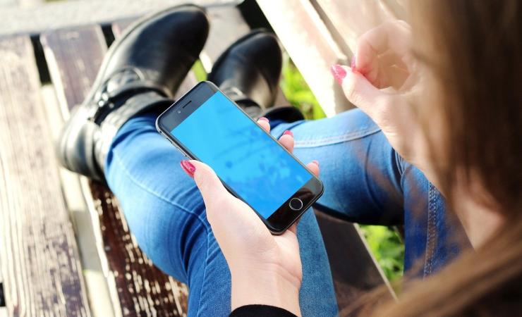 We are looking over the should of a girl sitting on what looks like a wooden bench. She's holding a smartphone in her left hand. She's wearing blue jeans and black ankle boots. Her skin is pale and her nails are painted a vivid pink colour.