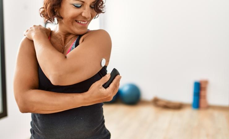 In the foreground of the picture is a woman with diabetes checking her glucose levels by scanning a glucose monitor attached to her upper arm. She is wearing sports clothes and in the background, out of focus, is some exercise equipment. Image by Krakenimages.com via Shutterstock