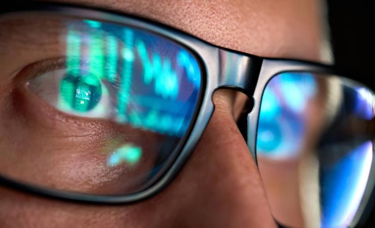 A close-up of the face of a man wearing dark-rimmed glasses. The glasses show the reflection of the screen he is looking at. Image by Ground Picture via Shutterstock.