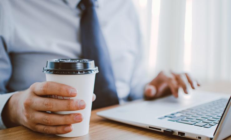 A man in a suit and tie looking at something on a laptop. He is holding a paper cup in his right hand. Image by Lisa Fotios via Pexels.