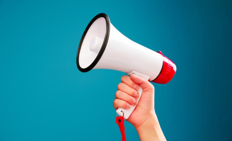 A picture of a hand holding up a megaphone against a turquoise background. Image by Sergey Mironov via Shutterstock