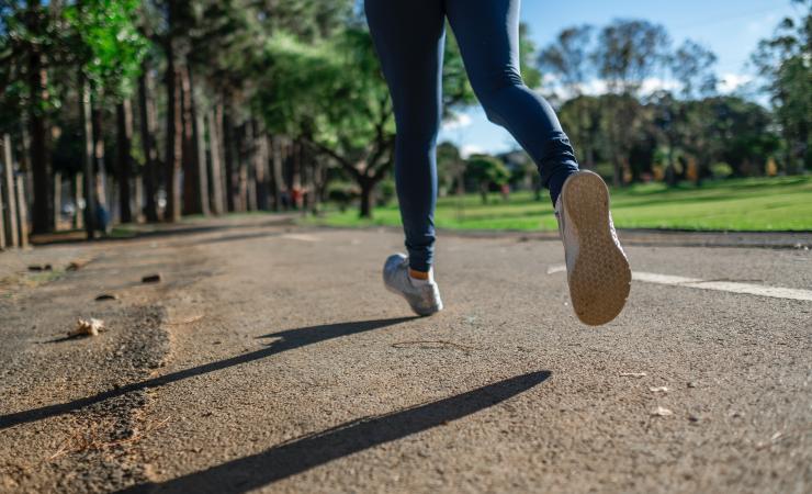 The legs of a person jogging. They're wearing blue leggings and trainers and they're jogging along a road with grass on the right and trees on the left. Image by Daniel Reche via Pexels