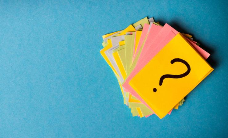 A pile of pieces of yellow, green, blue and pink paper against a blue background. The top piece of paper is bright yellow and has a question mark on it. Image by SNeG17 via Shutterstock.