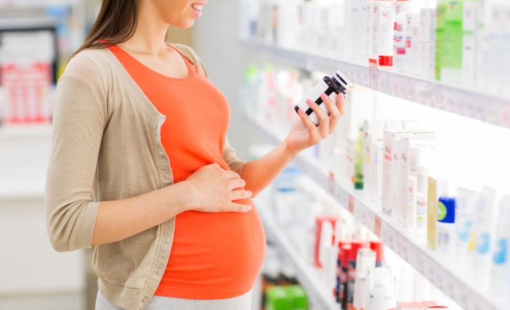 A pregnant woman in an orange t-shirt and beige cardigan looks at the label on a bottle of medicine. Her right hand rests on her bump. Image by Syda Productions via Shutterstock.
