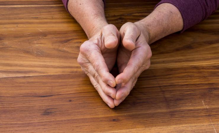 The hands of a man, held lightly together, resting on a wooden table top. The joints of the man's knuckles are visibly swollen. 