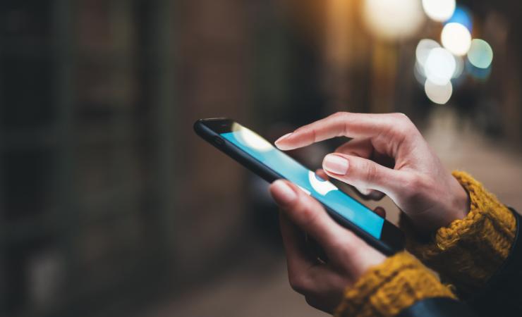 Close up of the hands of a woman using a smartphone. She has brown skin and is wearing a chunky knit ochre jumper. The background is blurred and quite dark. Image by A_B_C via Shutterstock.