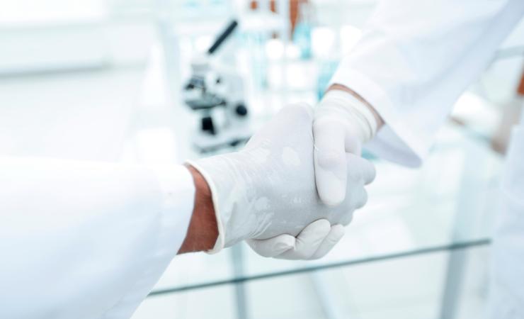 Two people in white labcoats and white rubber gloves are shaking hands. Image by ASDF_MEDIA via Shutterstock