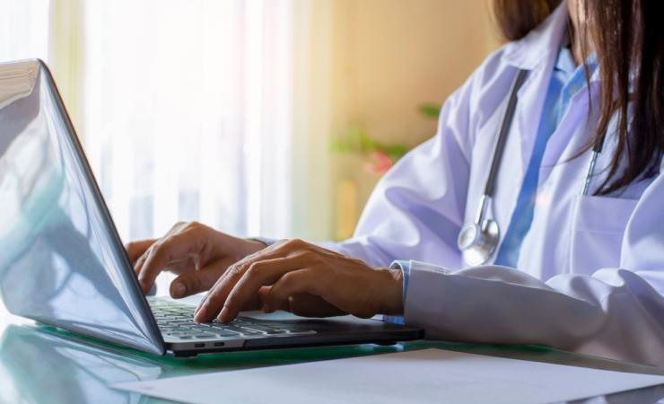 A woman doctor wearing a white coat and with a stethoscope around her neck typing at a laptop. Image by NIKCOA via Shutterstock.