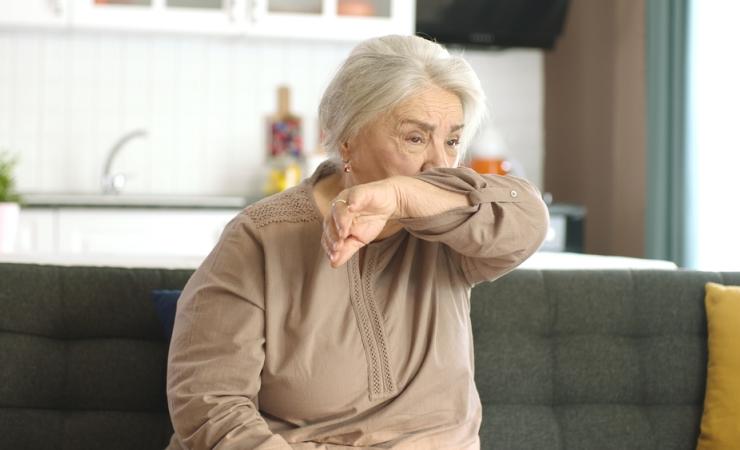 A senior woman coughing into her elbow. She has white skin and long white hair tied back. She's wearing a beige long-sleaved top and sitting on a grey sofa. Image by Red Stock via Shutterstock.