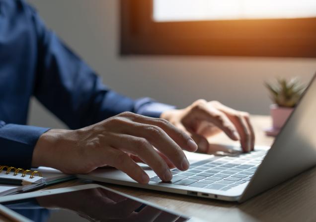 A man is typing on a laptop in front of a window. The laptop is open and the man's hands are on the keyboard. There are several notebooks and a tablet on the desk. Image by Thanadon88 via Shutterstock.