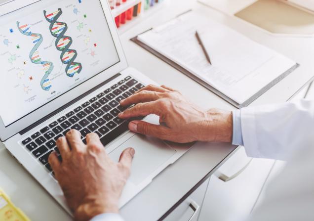 Scientist working at a laptop with a DNA double helix on the screen. Image by 4PM productions via Shutterstock.