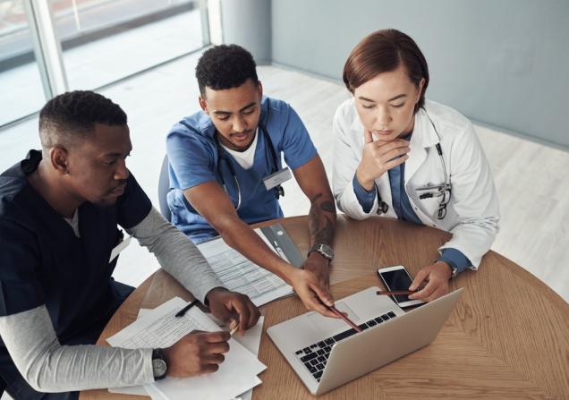 Three people sitting round a table looking at a laptop. Two of them have stethoscopes around their neck. Image by PeopleImages.com - Yuri A via Shutterstock.