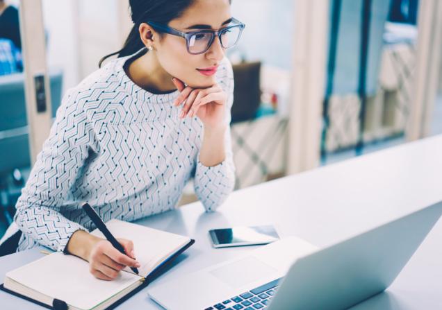 A woman looking at a computer screen and taking notes. Image by GaudiLab via Shutterstock