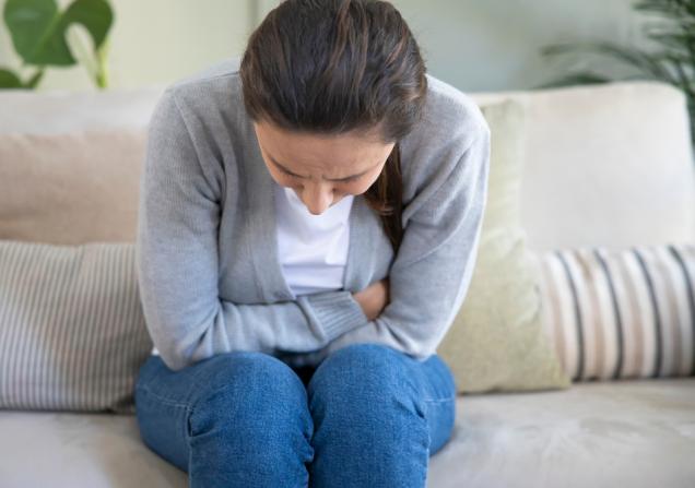 A woman is sitting on a sofa, facing towards us. She's doubled over in pain, so we can't really see her face, just the top of her head. She's wearing a white top, grey cardigan and blue jeans. Her long dark hair is tied back in a ponytail. Image by Ahmet Misirligul via Shutterstock.