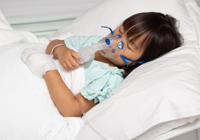 A young girl lying in a hospital bed with her eyes closed and an oxygen mask on. Image by Huttsu via Shutterstock.