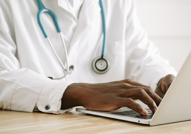 A close-up of a doctor typing at a computer. The doctor is wearing a white doctor's coat and a stethoscope. Image by Ivan Samkov via Pexels.