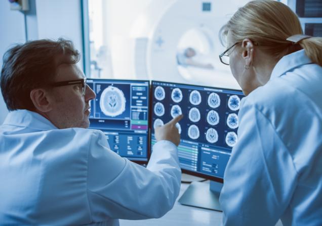 A doctor and radiologist discuss look at a monitor showing brain scan results, while a patient undergoes a scan in the background. Image by Gorodenkoff via Shutterstock.