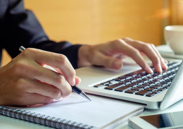 A person taking notes at a laptop. Image by mrmohock via Shutterstock