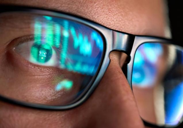 A close-up of the face of a man wearing dark-rimmed glasses. The glasses show the reflection of the screen he is looking at. Image by Ground Picture via Shutterstock.