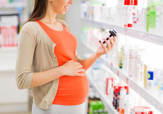 A pregnant woman in an orange t-shirt and beige cardigan looks at the label on a bottle of medicine. Her right hand rests on her bump. Image by Syda Productions via Shutterstock.