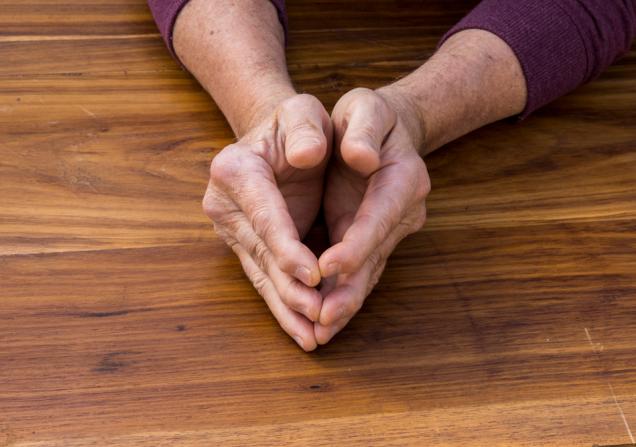 The hands of a man, held lightly together, resting on a wooden table top. The joints of the man's knuckles are visibly swollen. Image by Jacques Hugo via Shutterstock