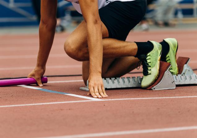 A sprinter at the start of a relay race. Their shoes are bright yellow/green and the baton is pink. Image by Real Sports Photos via Shutterstock