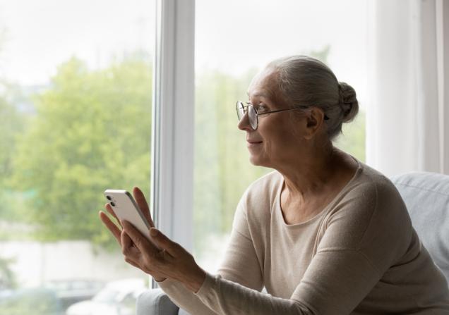 A senior lady using a smartphone. She has long grey hair tied back in a bun and glasses, and is wearing a light coloured sweater. She's sitting in front of a window. Image by fizkes via Shutterstock.