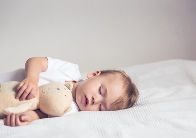 A baby is lying sleeping on its left side on a bed. The baby is cuddling a beige teddy bear. Image by Mallmo via Shutterstock.
