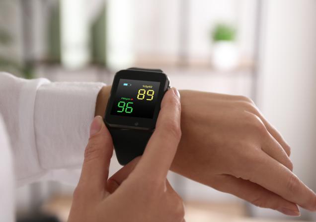 Close-up of a woman measuring oxygen levels with a smartwatch. The watch is on her left arm and she is holding the black watch with the thumb and index finger of her right hand. Image by New Africa via Shutterstock